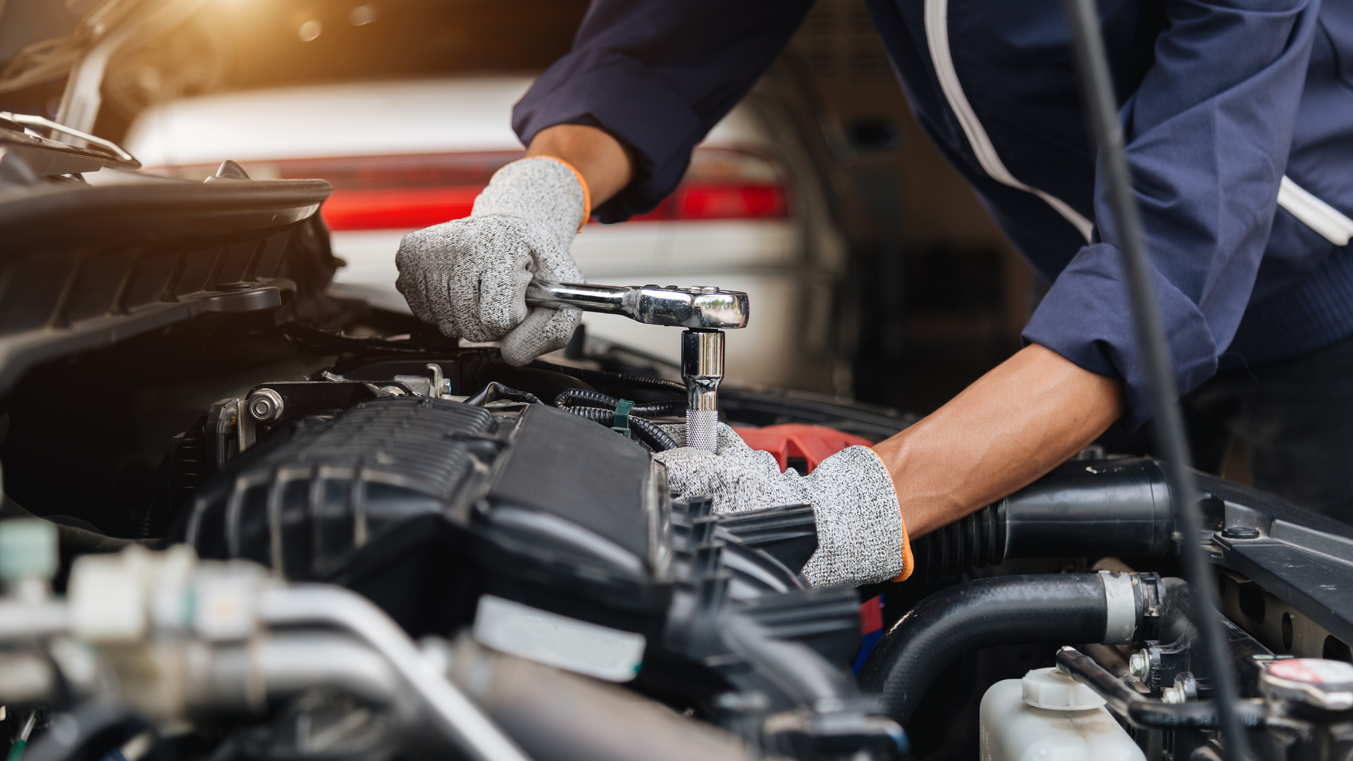 Mechanic in blue workwear uses a wrench on a car engine, wearing gloves. Outdoors, in daylight.
