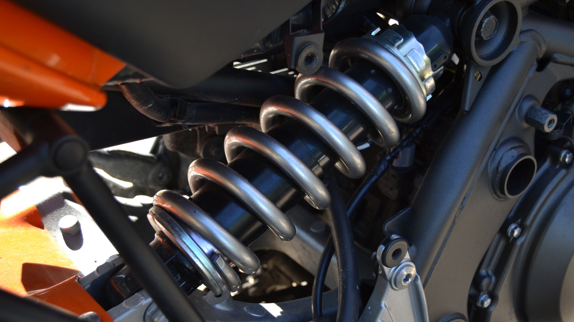 A close-up of a motorcycle's rear suspension system, featuring a silver coil spring, black frame, and orange components.