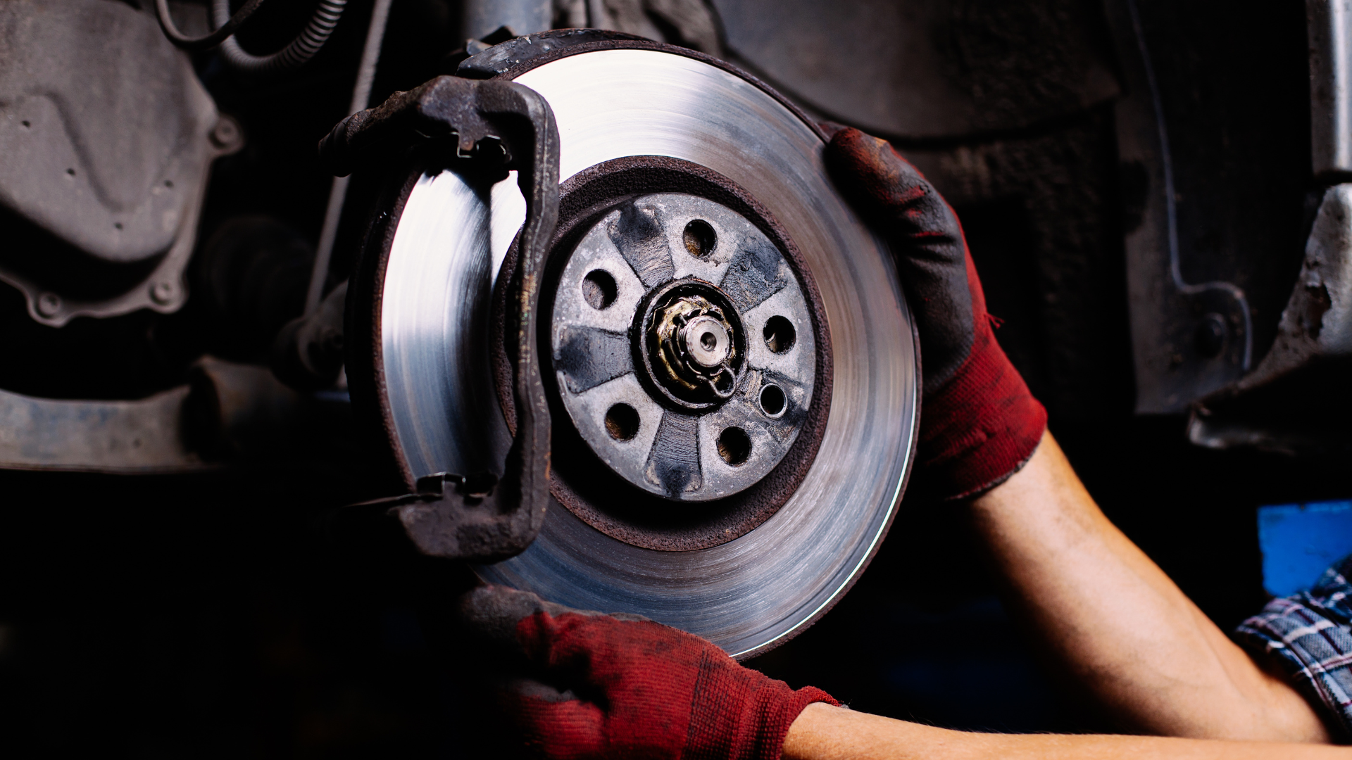 Person with gloved hands holding a car brake rotor and caliper. Red gloves, dark setting.