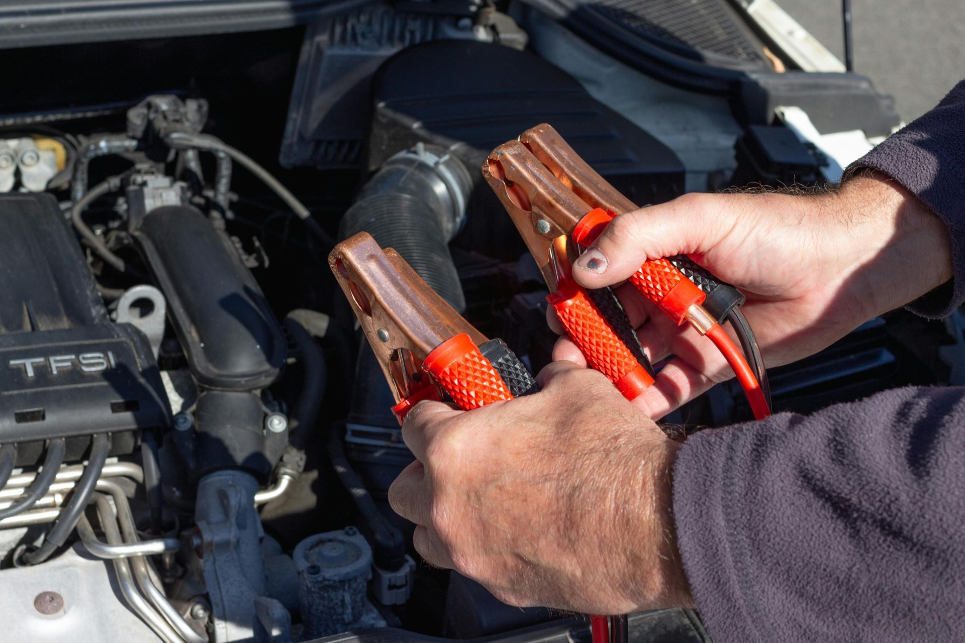 Hands holding red and black jumper cables near an open car hood.