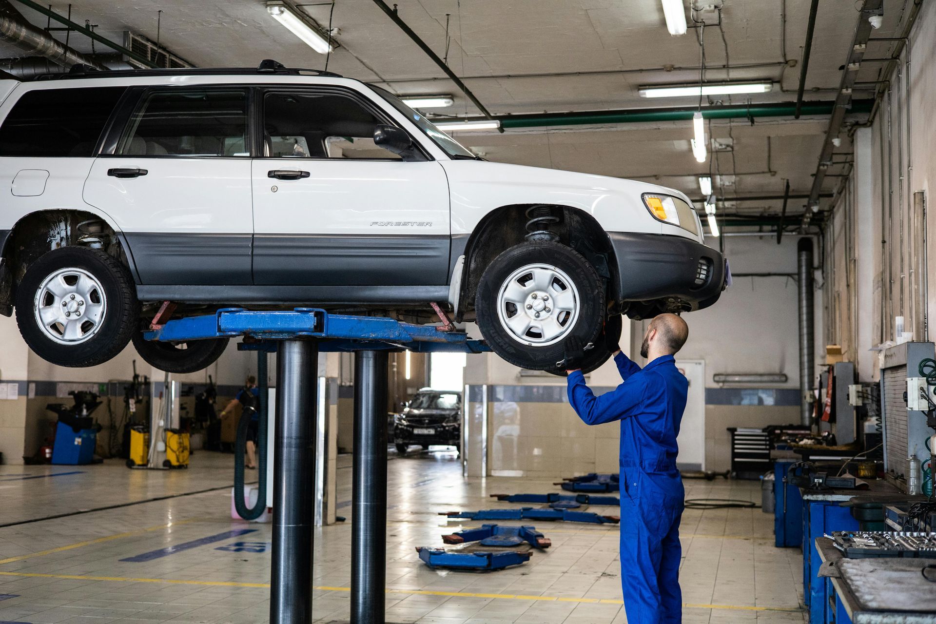 Mechanic in blue overalls working on white car raised on a lift in a garage.